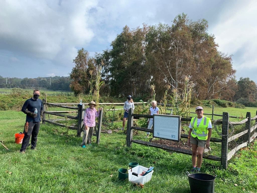 Several people stand near a small fenced in community garden in a green Connecticut field with trees in the background.